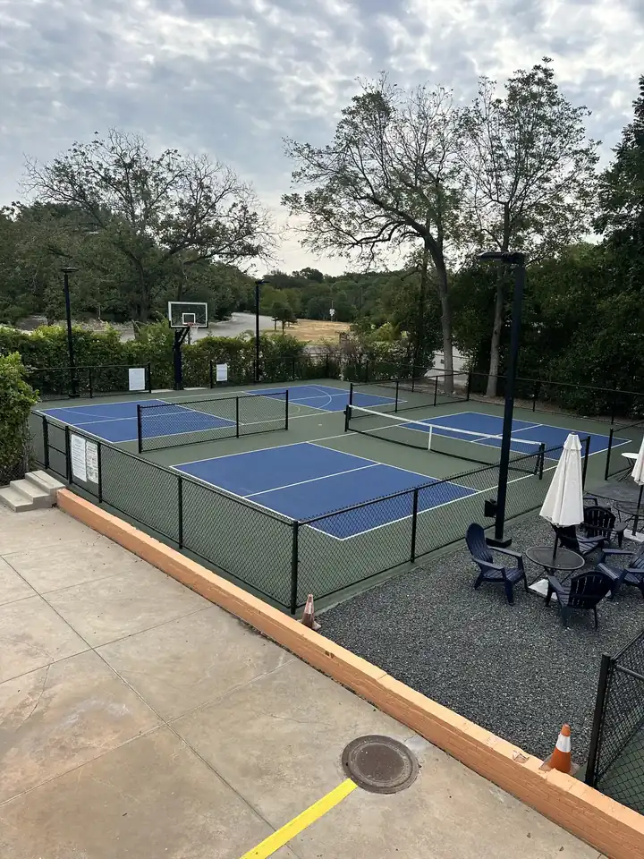Outdoor basketball hoop beside the fenced pickleball courts, umbrellas and chairs to the side