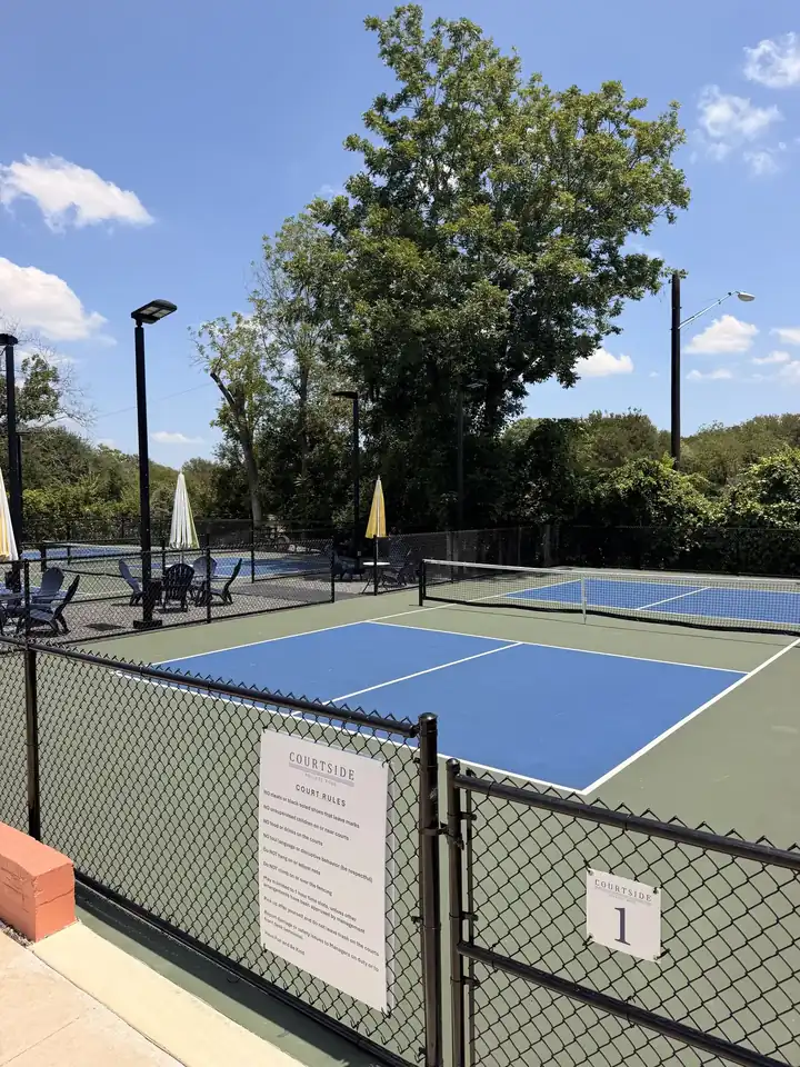 Blue pickleball courts behind a black chain-link fence with light poles and shade trees
