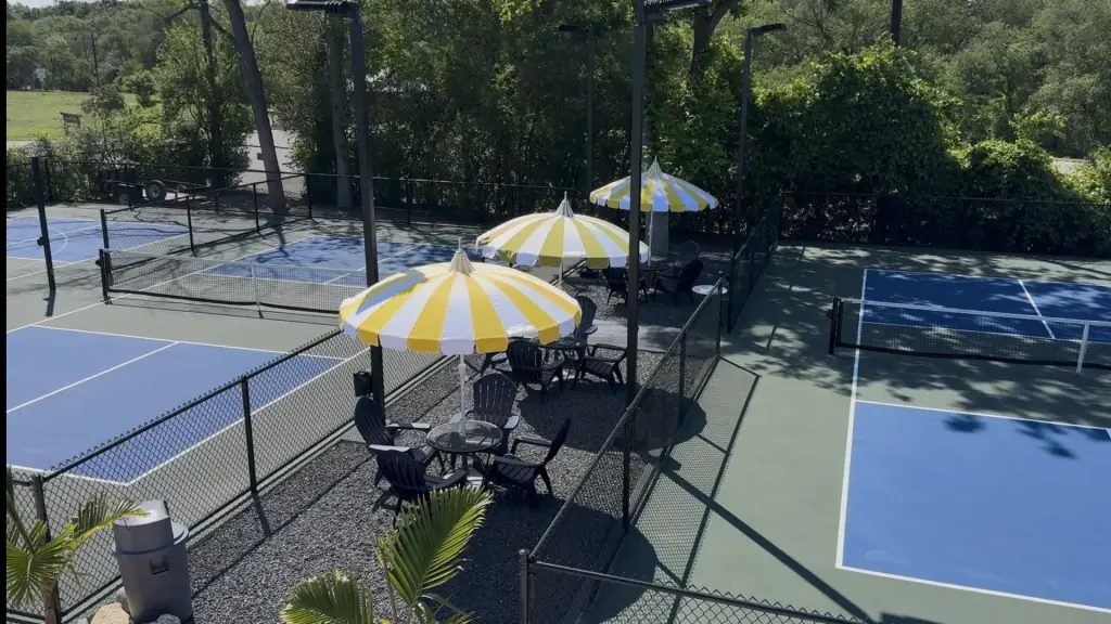 Blue pickleball courts in the foreground, yellow-and-white umbrellas and the pool deck beyond the fence