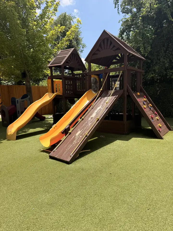 Wooden playscape with two yellow slides on green turf, trees behind